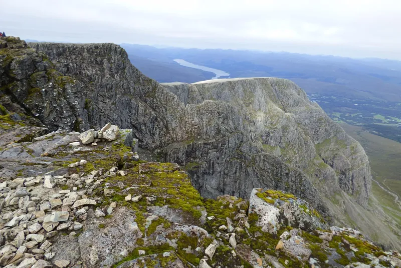 A rocky mountain ridge in the Scottish Highlands, looking down a steep crag to a loch in the glen below